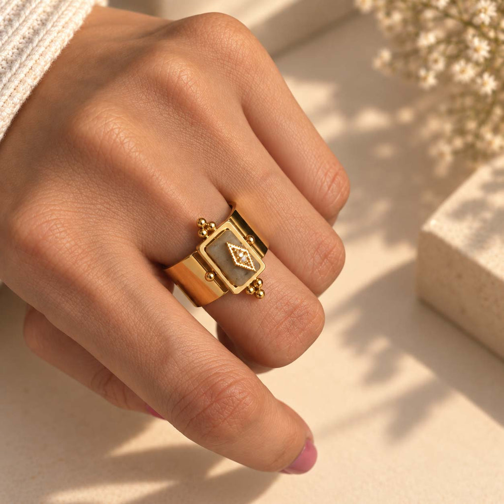 A Celeste Artisan Statement 14K gold plated ring with a rectangular face, a central gemstone motif, and small bead details, shown worn on a hand in warm sunlight.