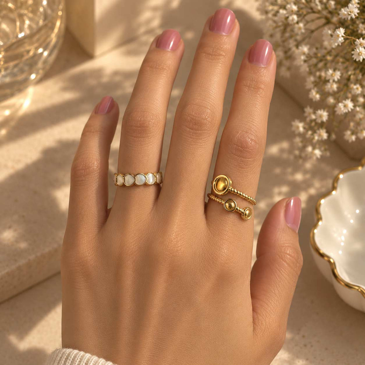 A close-up of a hand wearing two 18K gold rings, one with small white enamel inlays and the other with a round gold bead details, set against a warm, softly lit background with dried flowers.