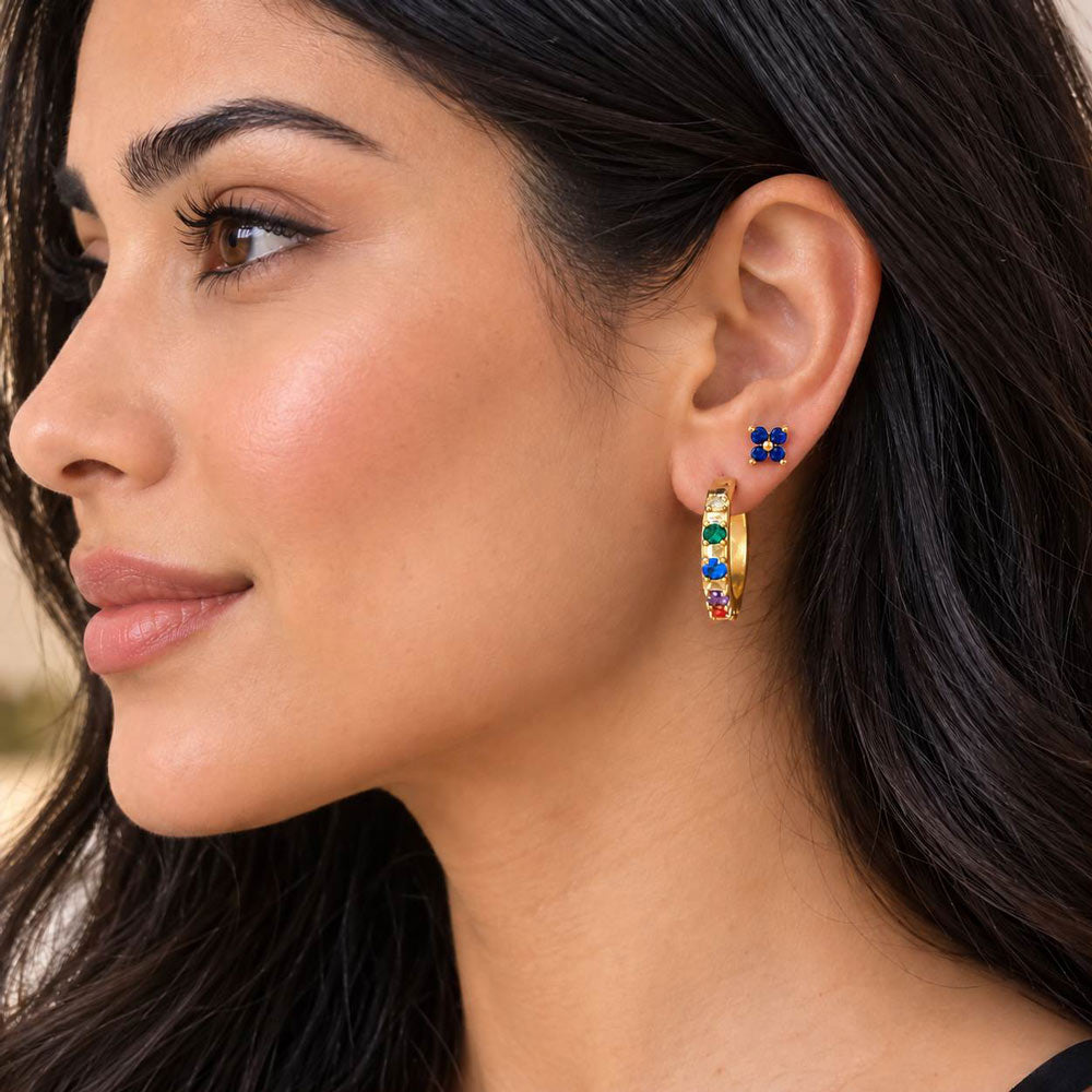 A close-up of a woman wearing a 18K gold plated huggie hoop earring adorned with multicolour gemstones paired with a small matching blue butterfly shaped helix stud earring.