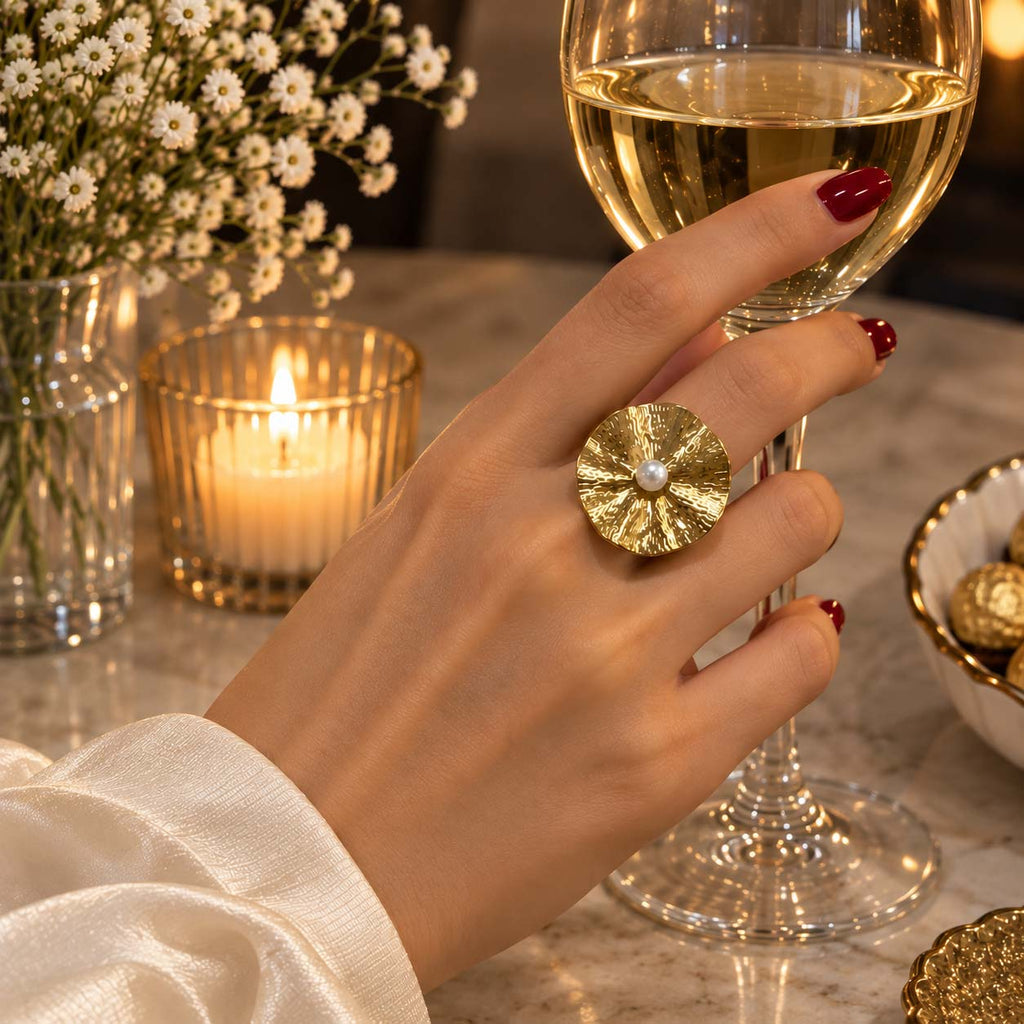 A hand wearing a large cocktail 18K gold plated ring with a textured sunburst pattern and a central white pearl, holding a glass of white wine in a warm, elegantly lit setting.