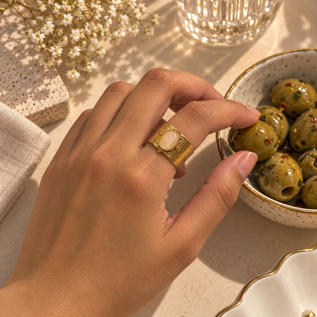 A close-up lifestyle shot of a hand wearing a 18K plated gold ring with a round gemstone in centre, reaching toward green olives in a speckled bowl on a warm, sunlit beige surface.
