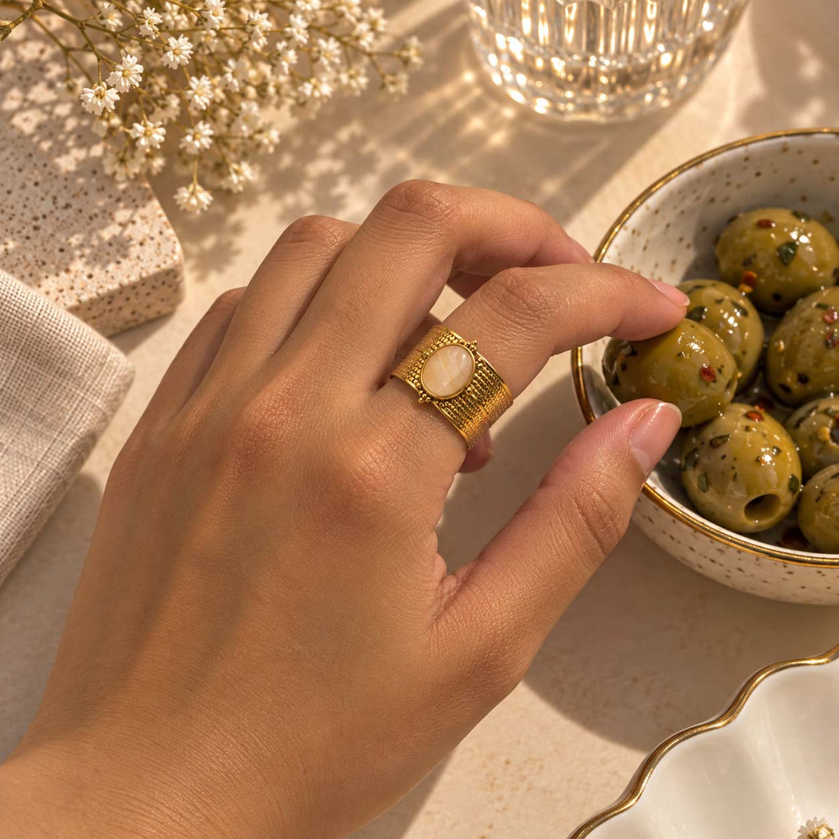 A close-up lifestyle shot of a hand wearing a 18K plated gold ring with a round gemstone in centre, reaching toward green olives in a speckled bowl on a warm, sunlit beige surface.