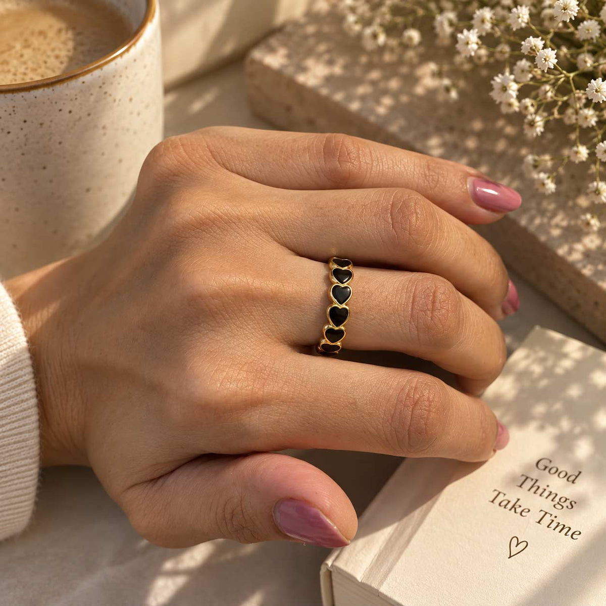 A close-up of a person's hand resting on a book, wearing a delicate 18K gold ring adorned with small black heart enamel inlays. 