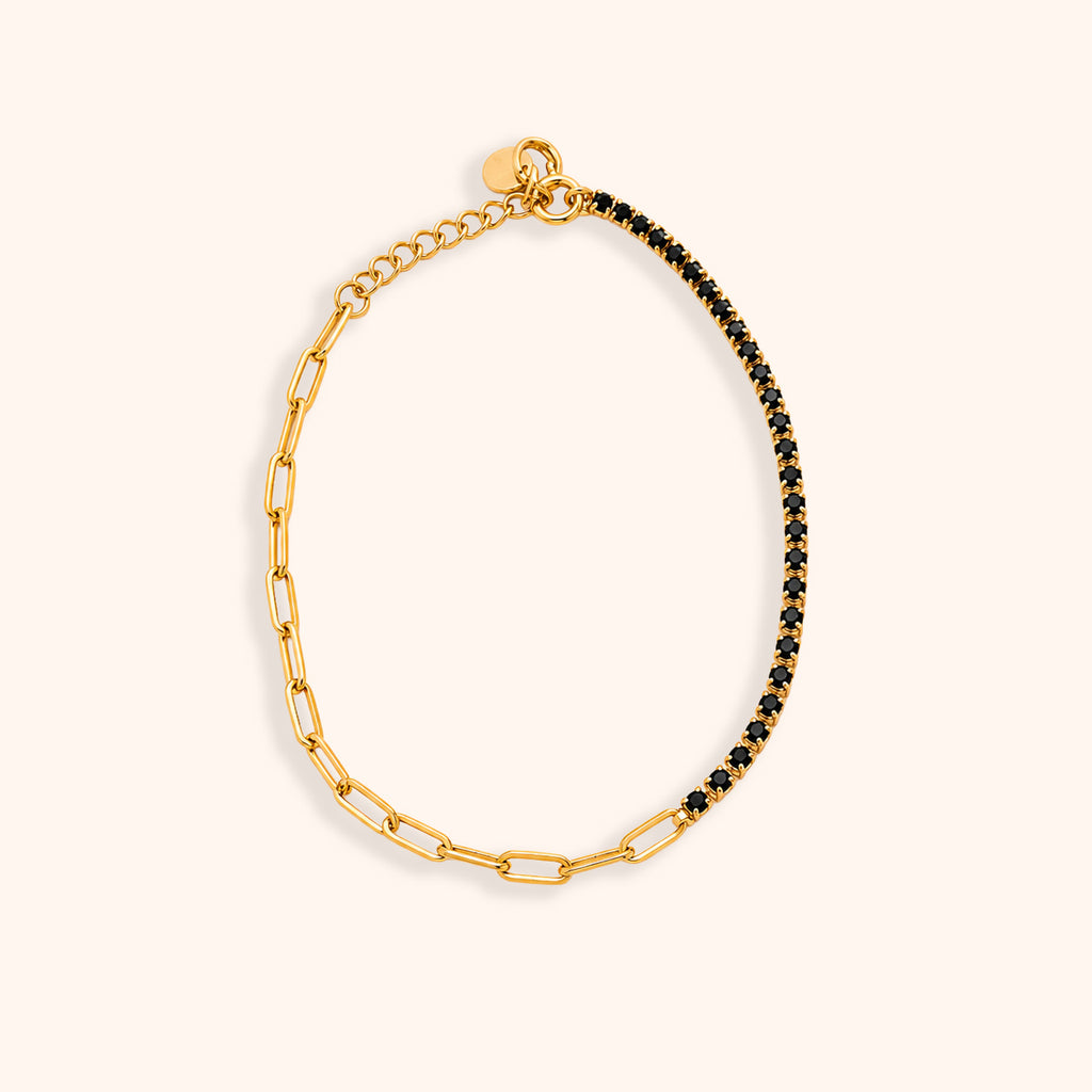 A gold paperclip chain bracelet with one half embellished with small black stones, displayed on a light cream background.