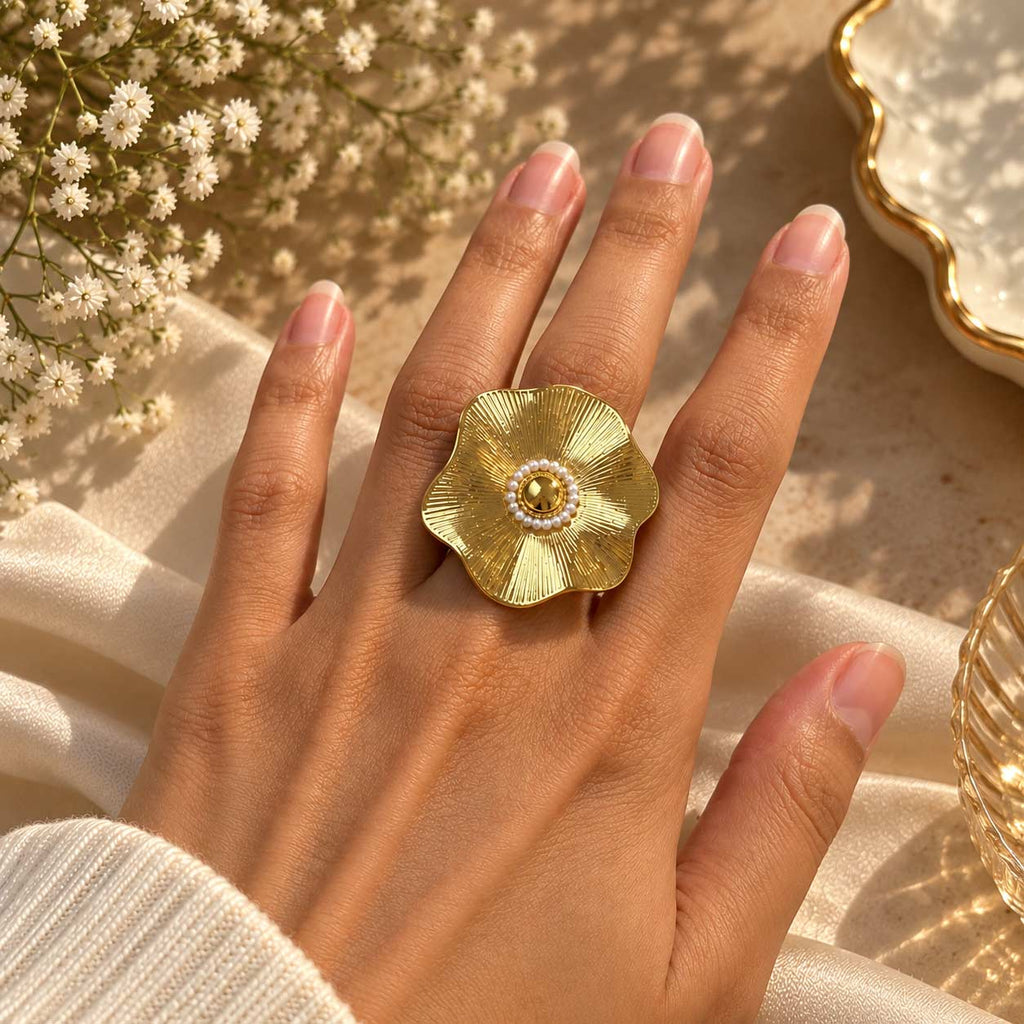 A gold flower-shaped ring with a pearl center, worn on a hand resting on draped cream fabric with small white flowers in the background.