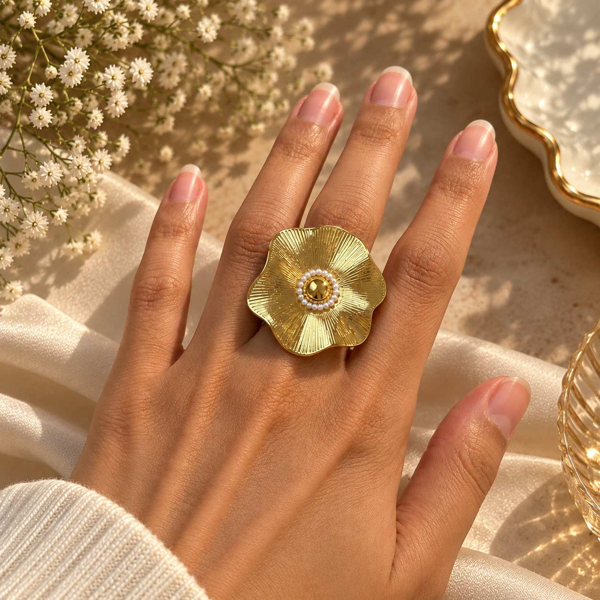 A gold flower-shaped ring with a pearl center, worn on a hand resting on draped cream fabric with small white flowers in the background.