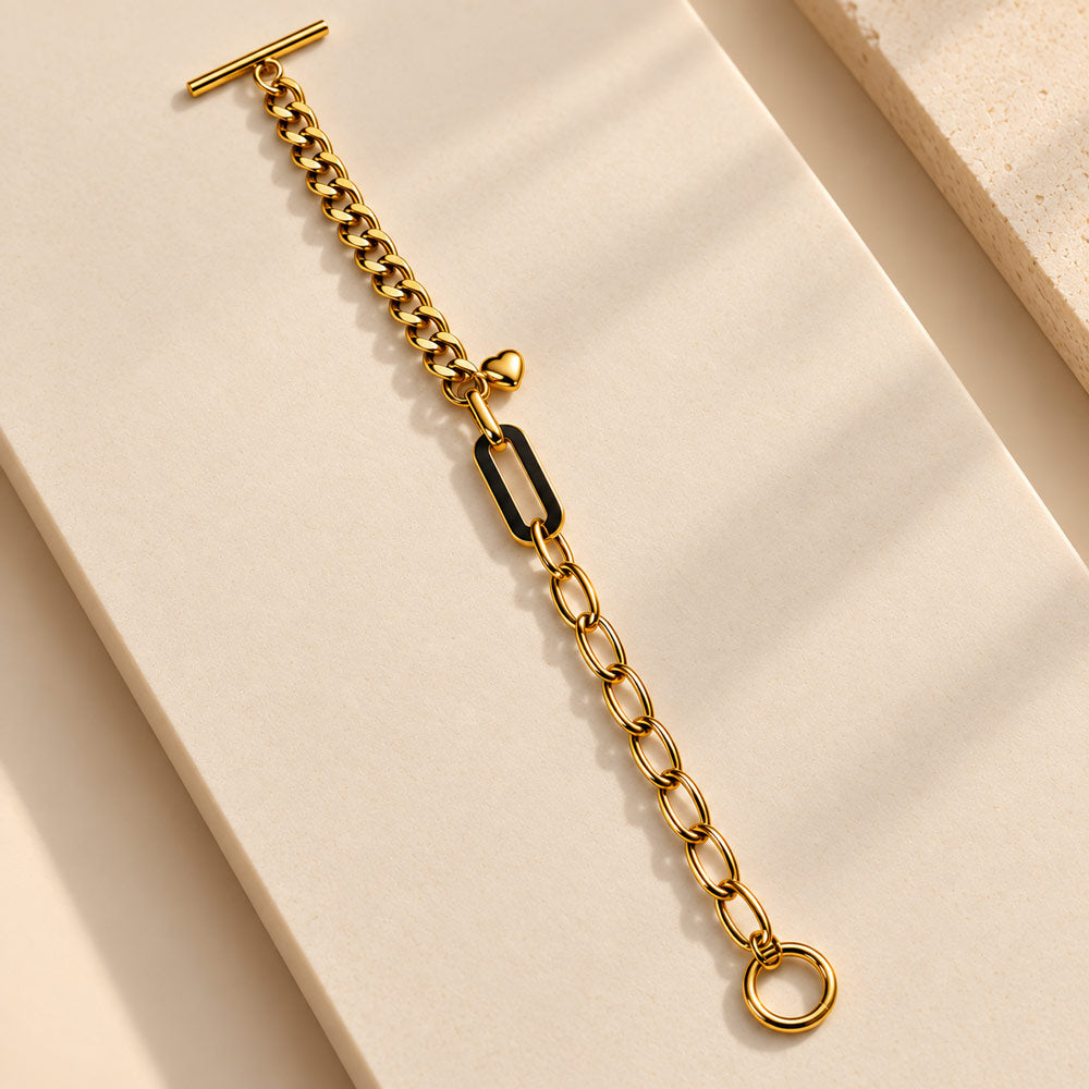 A gold chain bracelet with a black rectangular link accent and a T-bar clasp, displayed on a warm beige stone surface.
