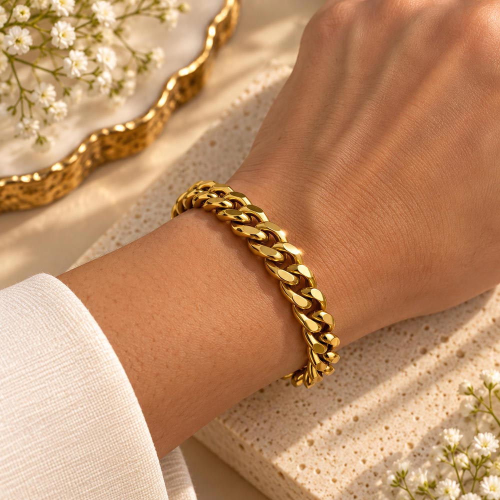 A close-up of a person's wrist wearing a polished gold Cuban link chain bracelet, styled with a textured beige stone slab and delicate white flowers.