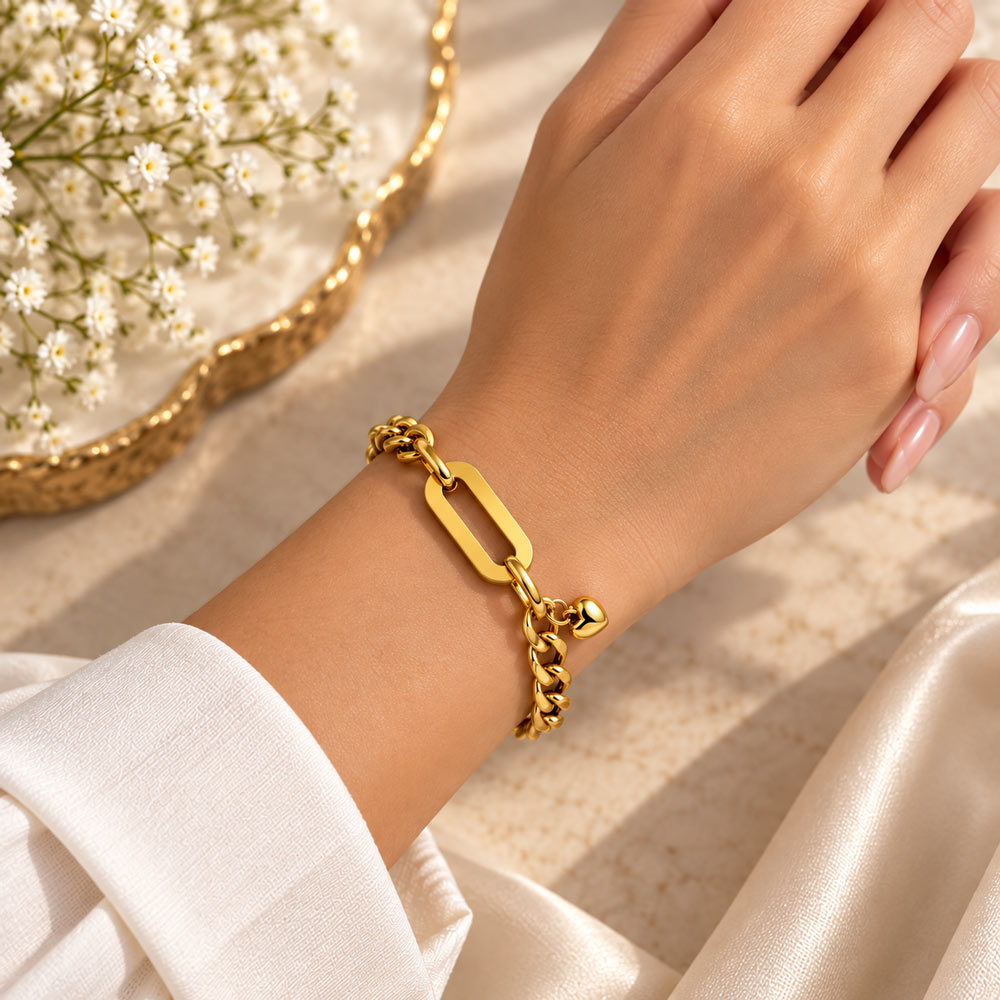 A close-up of a person's wrist wearing a polished gold chain bracelet with a rectangular link and small dangling charm, styled with white satin fabric and baby's breath flowers.