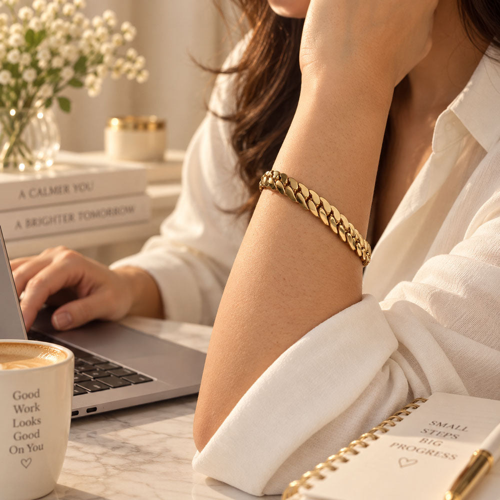 A close-up lifestyle shot of a woman wearing a chunky gold chain link bracelet on her wrist, sitting at a desk with a laptop, coffee, and a notebook.