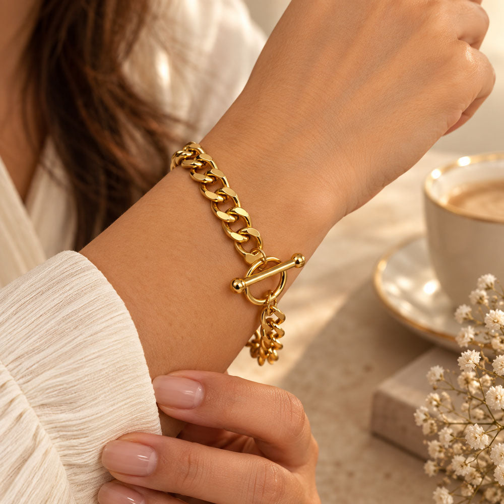 A close-up lifestyle shot of a woman's wrist wearing a chunky gold Cuban link bracelet with a toggle clasp and a dangling leaf charm. The background features a soft-focus coffee cup and dried baby's breath flowers.