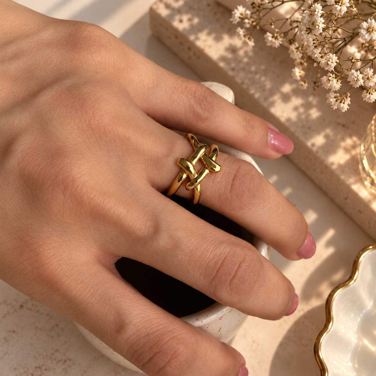 A close-up of a hand wearing a 18K gold plated ring with an interlocking knot design, resting on a white ceramic cup with dried flowers in the background.