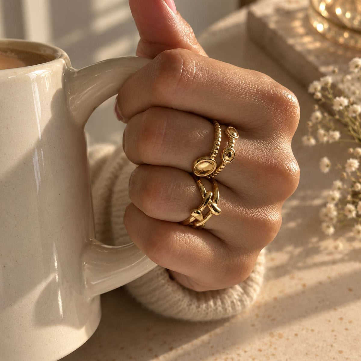 A close-up of a hand wearing two gold rings. The 18K gold-plated rings feature organic, textured gold designs, and the background includes a cozy knit sleeve and small dried flowers.