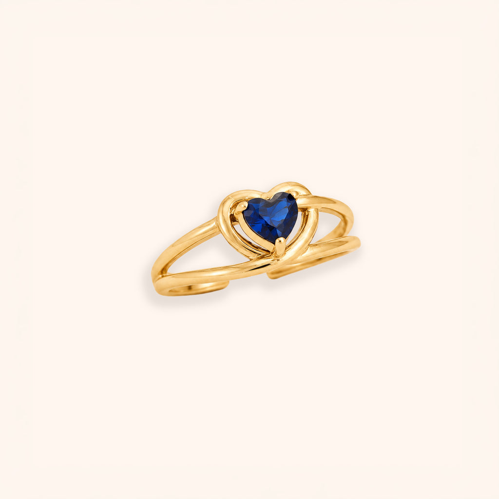 A 18K gold-plated open ring band featuring a heart-shaped dark blue gemstone at the center, set against a solid off-white background.