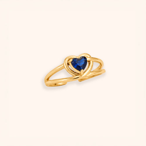 A 18K gold-plated open ring band featuring a heart-shaped dark blue gemstone at the center, set against a solid off-white background.