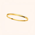 A minimalist, polished yellow gold bangle bracelet shown on a clean, solid, warm off-white background.