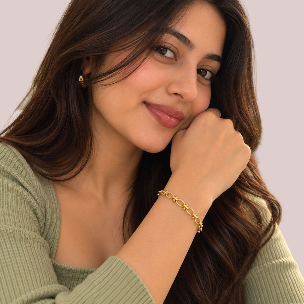A woman with long dark brown hair wearing a delicate gold chain bracelet on her wrist, resting her chin on her hand against a soft, light lavender background.