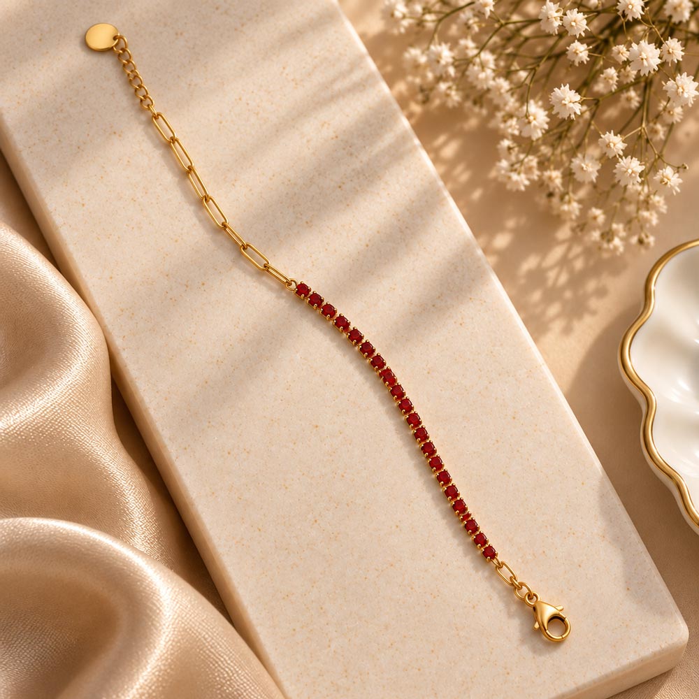 A gold chain bracelet with a central row of small red gemstones, displayed diagonally on a textured cream stone block with beige satin fabric and white baby's breath flowers.