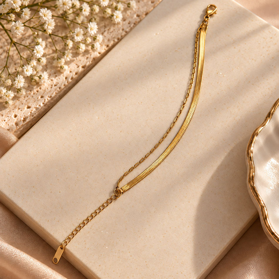 A gold snake chain bracelet with a lobster clasp and extender chain, styled on a beige textured book with dried baby's breath flowers and a gold-rimmed dish.