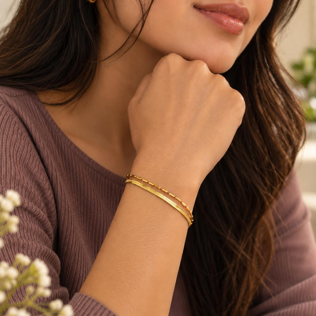 A close-up lifestyle shot of a woman wearing two delicate gold bangles on her wrist, wearing a mauve ribbed knit top.
