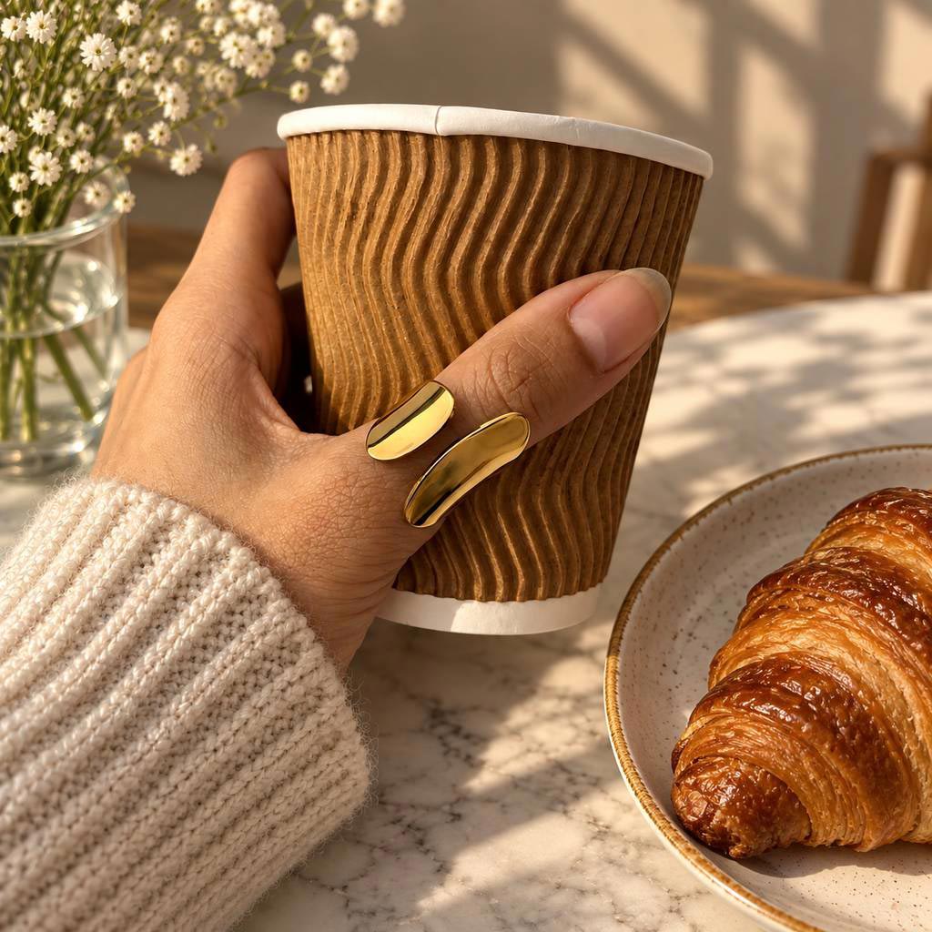 A close-up lifestyle shot of a hand holding a brown corrugated paper coffee cup while wearing 18K gold plated ring on the thumb. 