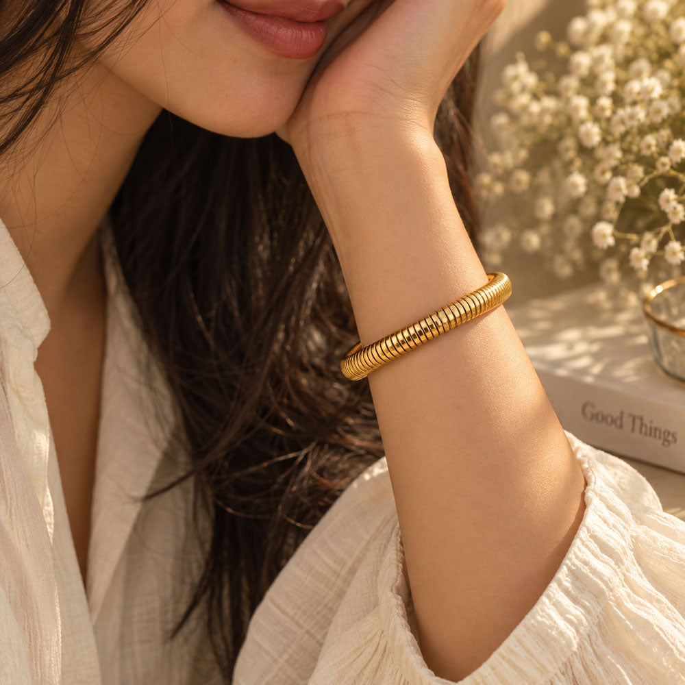 A close-up lifestyle shot of a woman wearing a textured 18K gold plated bangle bracelet on her wrist, resting her head on her hand in warm, sunlit surroundings.