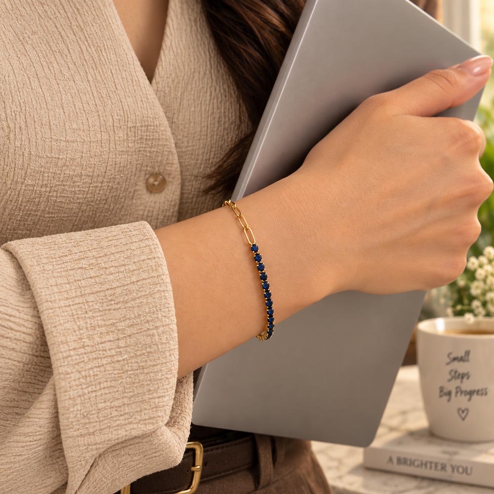 A close-up lifestyle shot of a woman's wrist wearing a delicate gold chain bracelet with small dark blue stones, while she holds a gray notebook in a softly lit indoor setting.