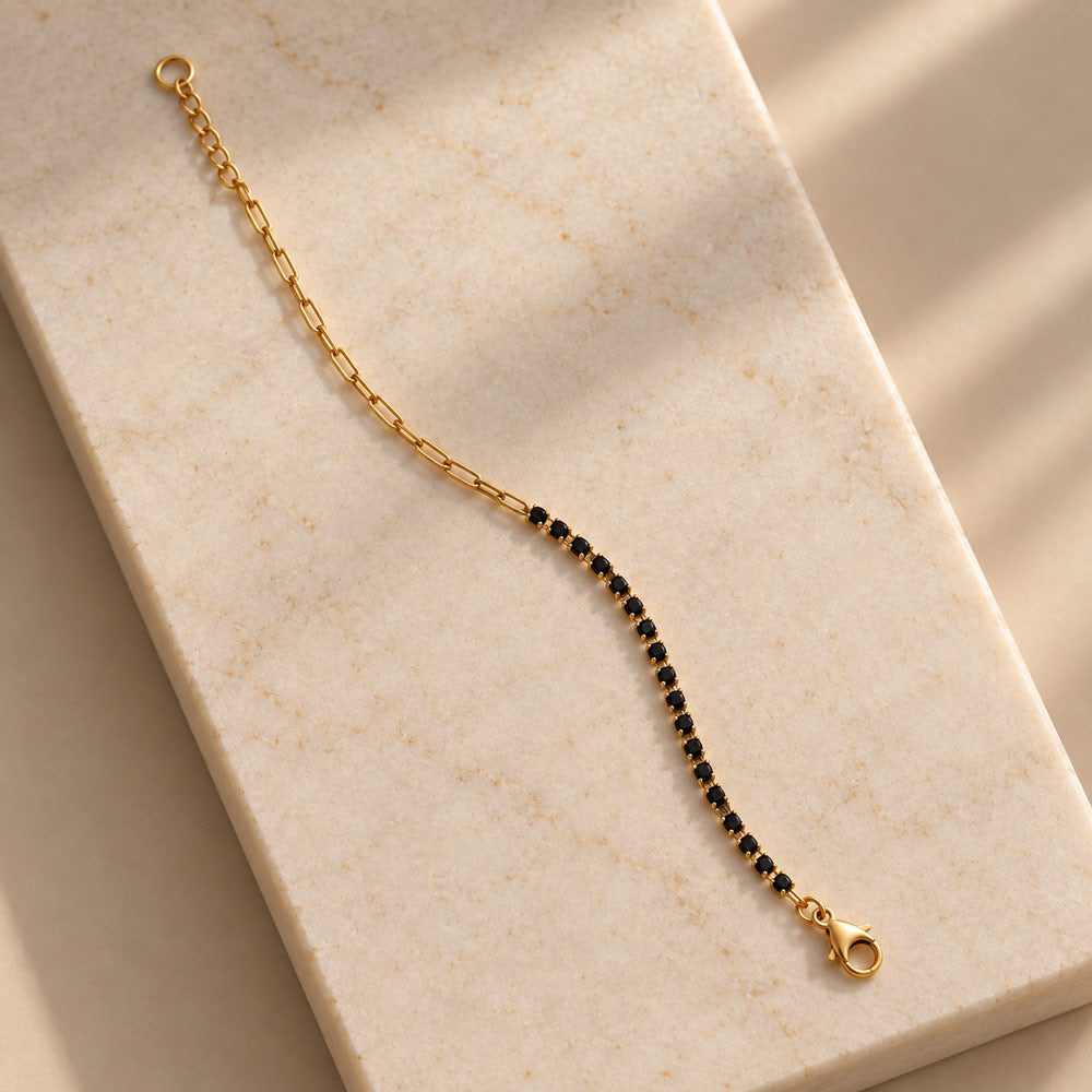 A delicate gold chain bracelet with a line of small black stones, displayed on a beige marble block with soft shadows.