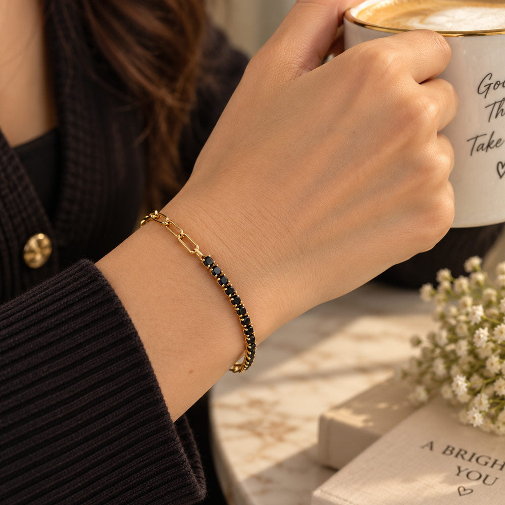 A close-up lifestyle shot of a person's wrist wearing a delicate gold chain bracelet with small dark blue stones, while holding a white coffee mug in a warm indoor setting.