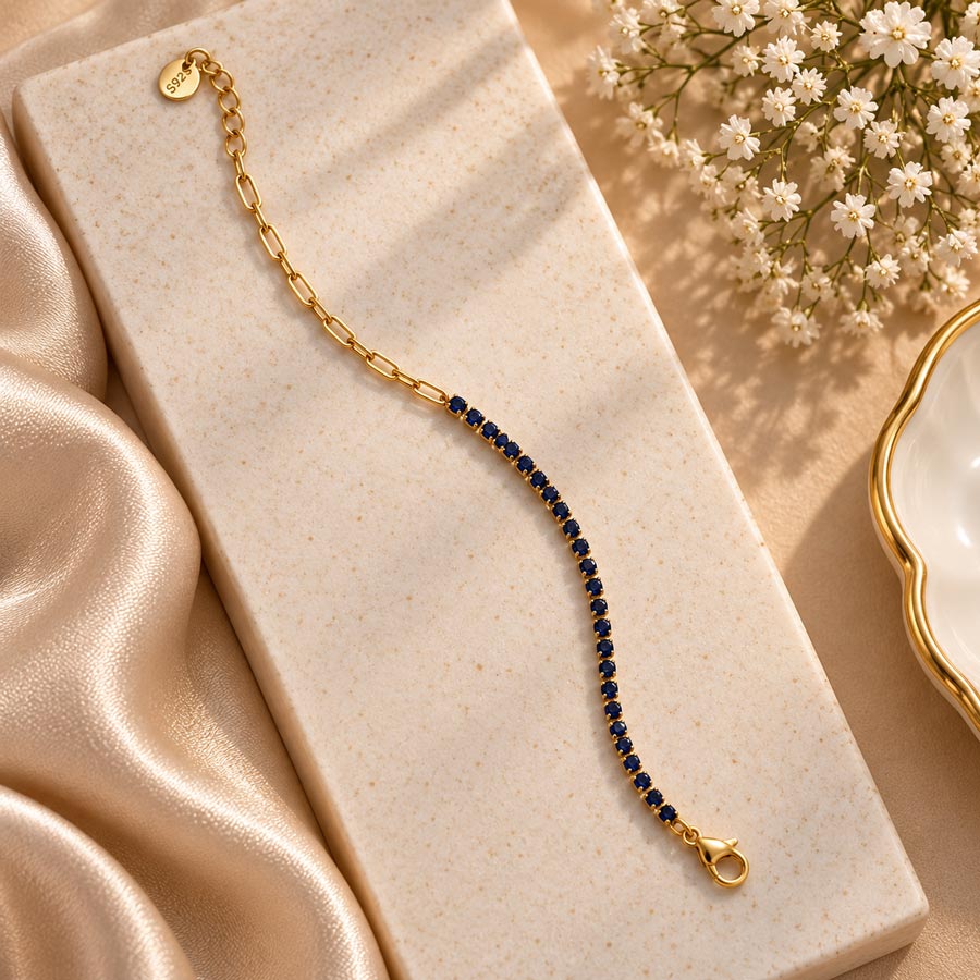 A gold chain bracelet with a single row of dark blue gemstones, displayed on a cream-colored stone block with gold satin fabric and white baby's breath flowers.