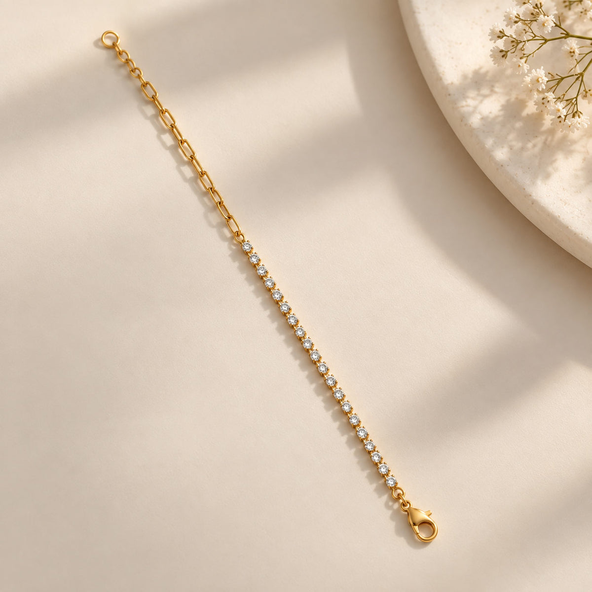 A gold tennis-style chain bracelet with a row of clear stones, displayed diagonally on a warm beige surface with soft shadows and a decorative plate corner in the background.