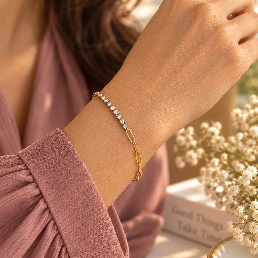 A close-up of a woman's wrist wearing a delicate gold chain bracelet with a row of clear rhinestones. She is wearing a dusty rose long-sleeve blouse, with a soft-focus background featuring white baby's breath flowers and a book.