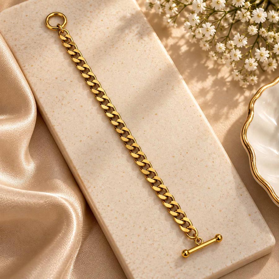 A gold-tone chain bracelet with a toggle clasp, displayed diagonally on a speckled cream stone slab with champagne satin fabric and small white flowers in the background.