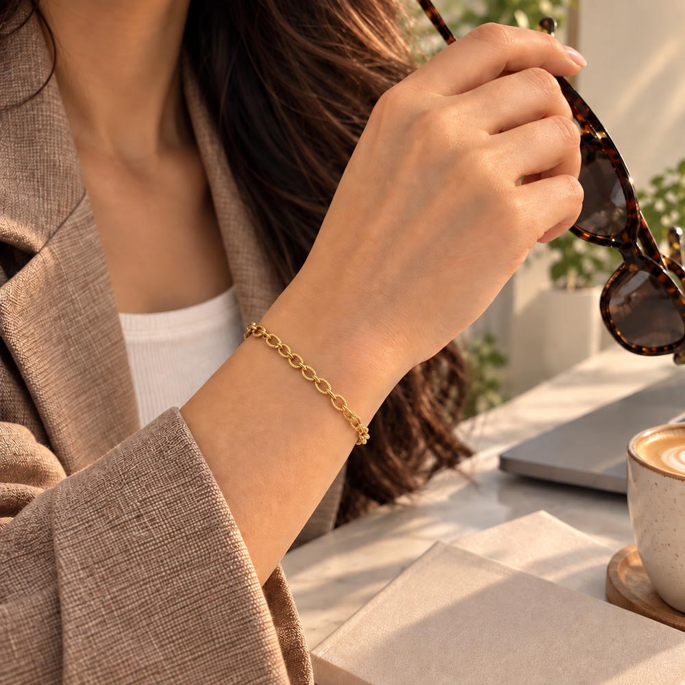 A close-up lifestyle shot of a woman wearing a delicate gold chain bracelet on her wrist, holding tortoiseshell sunglasses in a bright, modern indoor setting.