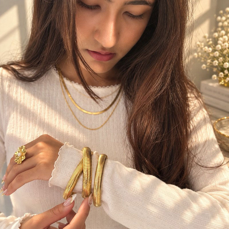 A woman wearing textured 18K gold bangle bracelets on her wrist, styled with layered gold necklaces and a gold ring, in a brightly lit indoor setting.