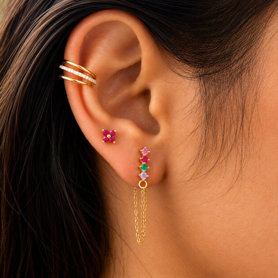 Close-up of a person's ear showcasing a gold ear cuff with small clear stones and a matching gold stud earring with pink, green, and purple gemstones and a dangling chain.