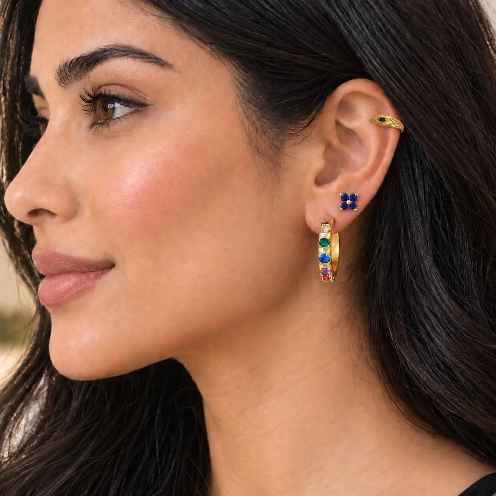 Close-up of a woman wearing a gold huggie hoop earring adorned with colorful gemstones in a rainbow sequence, along with a matching gold plated serpent ear cuff.