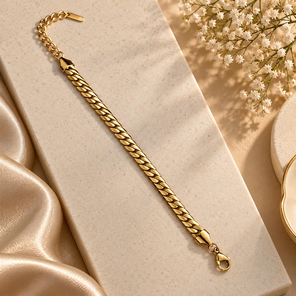 A gold-toned Cuban link chain bracelet styled on a textured cream rectangular block with champagne satin fabric and small white flowers in the background.